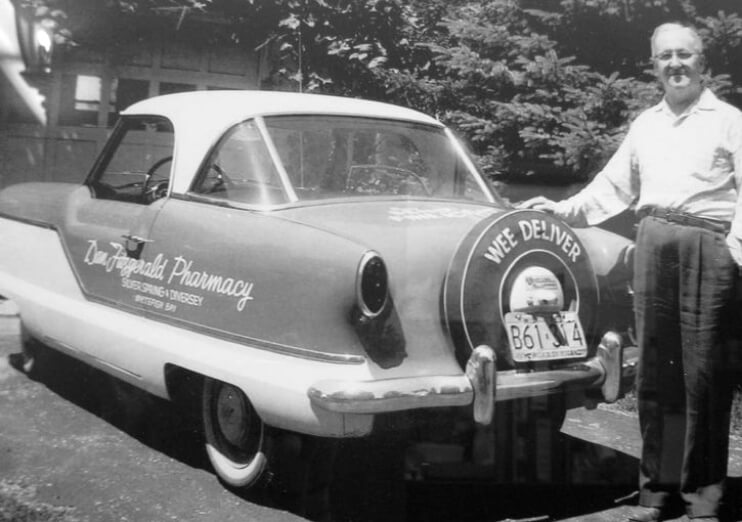 Black and white photo of Dan Fitzgerald standing next to a 1950s car, with the pharmacy's signage and message "Wee Deliver"