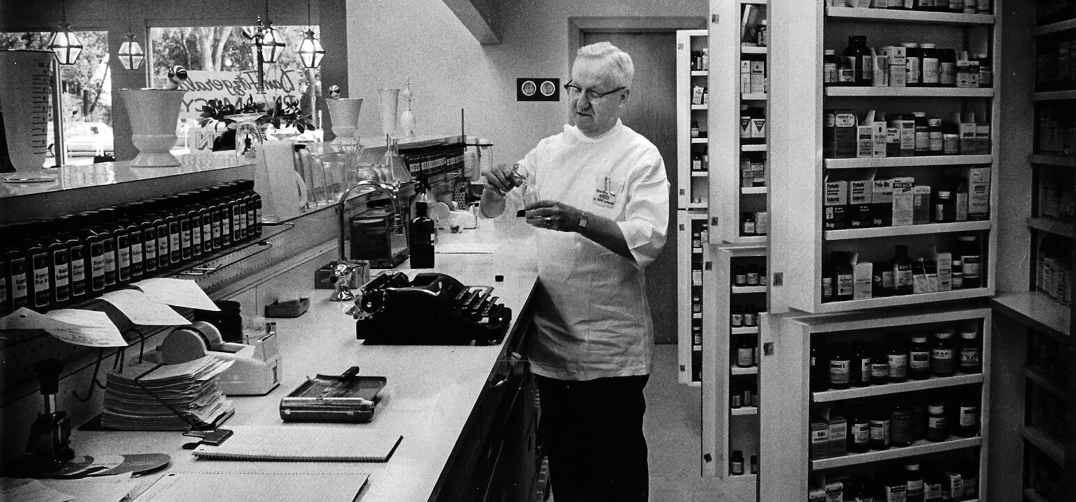 Black and white photo of Dan Fitzgerald behind the pharmacy counter, measuring pharmaceuticals